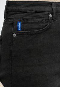 Nærbillede af lomme på sorte denimjeans med blå stofmærke mærket "HUGO" og synlige metalnitter i lommens hjørner.