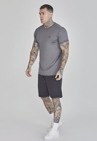 Tricou athletic gri cu mâneci scurte și design ajustat, purtat cu pantaloni scurți negri. Tenisi albi și tatuaje vizibile pe brațe și picioare.