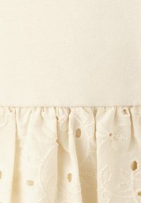 Tissu crème avec une section supérieure lisse et une section inférieure froncée brodée comportant des motifs floraux et des découpes circulaires.