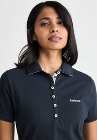 Ναυτικό πόλο μπλουζάκι με κοντά μανίκια, υφασμάτινη υφή, κουμπωτή στήθους και καρώ σχέδιο στον γιακά. Διαθέτει κεντημένο λογότυπο.