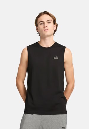 Giovane uomo con le braccia dietro la schiena che indossa una canotta nera senza maniche e pantaloncini grigi su uno sfondo chiaro uniforme.