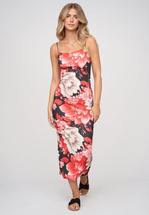 Junge Frau mit langen blonden Haaren, die ein schwarzes Kleid mit großem roten und beigem Blumenmuster und schwarze Sandalen trägt und vorwärts geht.