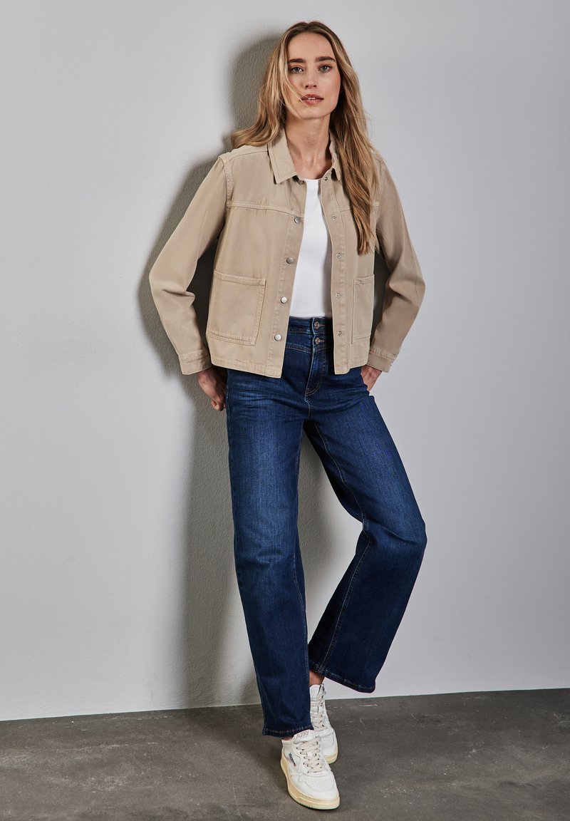 Veste en toile beige avec fermeture à boutons et deux poches avant, assortie à un jean droit bleu foncé et des baskets blanches. Texture lisse.
