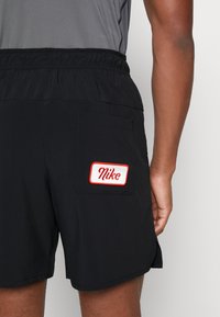 Shorts noirs avec une ceinture élastique, dotés d'une poche arrière avec un patch blanc et le texte "Nike" en rouge. Tissu lisse et coupe jusqu'aux genoux.