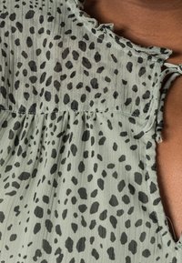 Blouse verte avec un motif à taches noires, présentant un col à volants et une texture plissée. Le tissu semble léger et fluide.