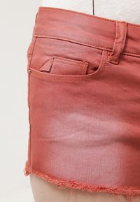 Lähikuva haalistuneesta punaisesta farkkuhameesta, jonka helma on revitty ja etutasku näyttää farkkutyylisiä ompeleita sekä vyölenkkiä.