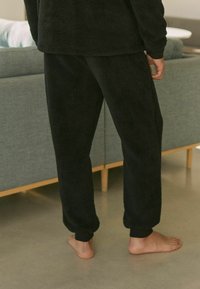Fekete fleece pizsama nadrág, amely puha tapintású, rugalmas mandzsettákkal és laza szabással rendelkezik. Meztelen lábbal viselve, világos padlón.