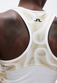 Κοντινή εικόνα του άνω μέρους της πλάτης ενός ατόμου που φοράει αθλητική μπλούζα τύπου racerback σε μπεζ και λευκό με ένα μικρό μαύρο λογότυπο κοντά στη γραμμή του λαιμού.