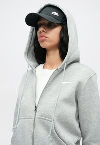 Sieviete ģērbusies pelēkā Nike kapucē ar rāvējslēdzēju un uzliktu kapuci, ar melno Nike vizieri, nedaudz skatoties uz sāniem pret vienkāršu fonu.