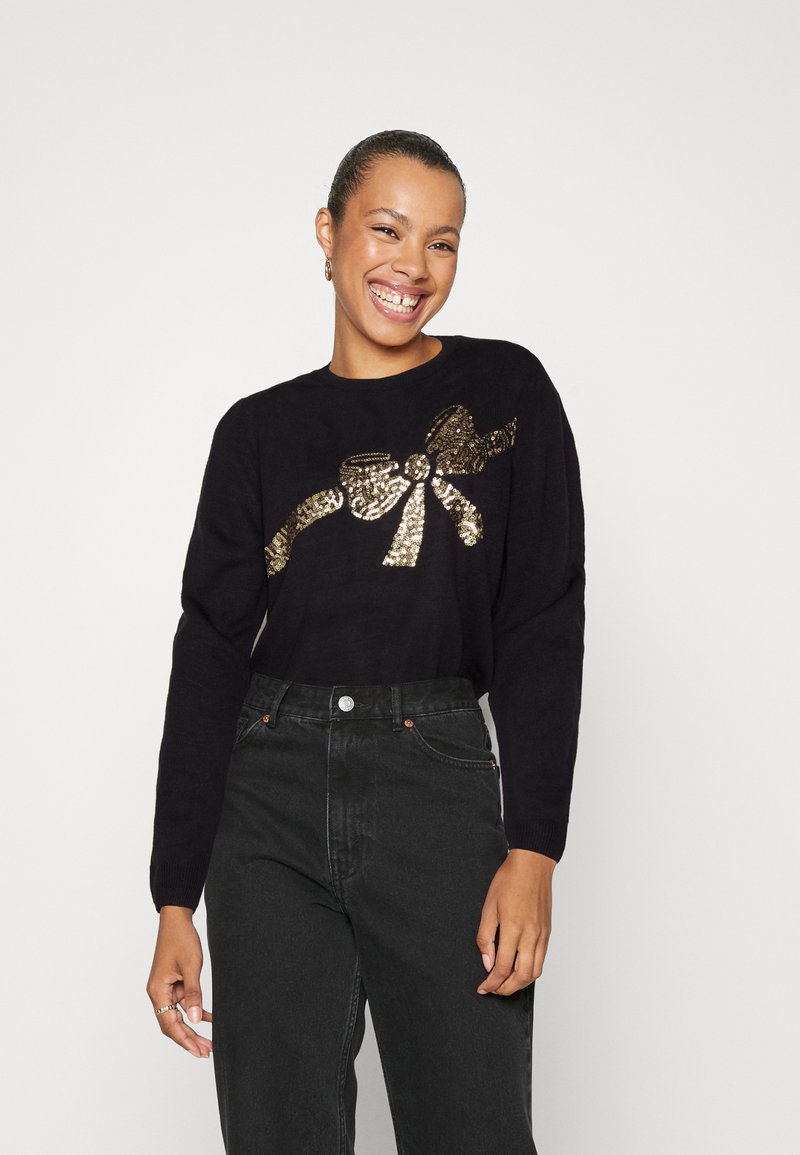 Pull noir avec un design de nœud en sequins dorés, associé à un jean noir taille haute. Le tissu semble doux avec une coupe décontractée.