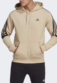 Hombre vistiendo una sudadera con cremallera beige de Adidas con rayas negras en las mangas y pantalones negros de Adidas con el logo en el muslo izquierdo.