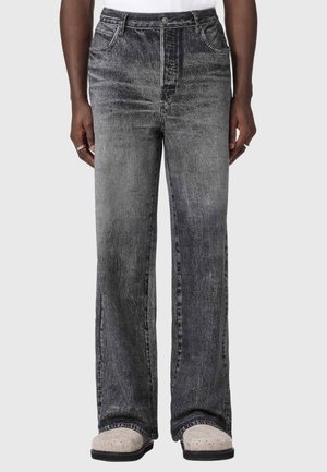 Person trägt ausgewaschene schwarze Jeans mit weitem Bein und helle Schuhe, steht mit entspannten Händen an den Seiten.