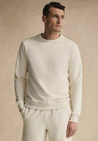 Krämfärgad sweatshirt med rund hals och lång ärm. Tillverkad av mjukt tyg, har ribbade muddarna och nederkant, ihop med matchande byxor.