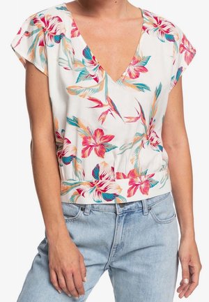 Frau trägt eine weiße V-Ausschnitt-Bluse mit pinkem, orangefarbenem und türkisfarbenem Blumenmuster, kombiniert mit hellblauen Jeans.