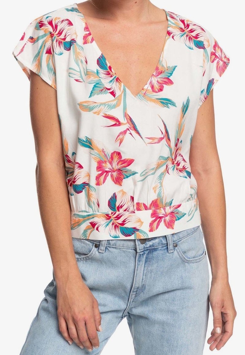 Femme portant un chemisier blanc col en V avec un imprimé floral rose, orange et bleu sarcelle, associé à un jean denim bleu clair.