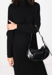 Robe col roulé côtelée noire à manches longues. Tient un sac bandoulière en cuir noir avec une poche zippée et des accents cloutés.