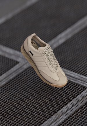 Beige sneakers med en glat læderoverflade, kontrasterende mørke snørebånd og en gummisål. Tre-stribet design på siden og mærket på tungen.