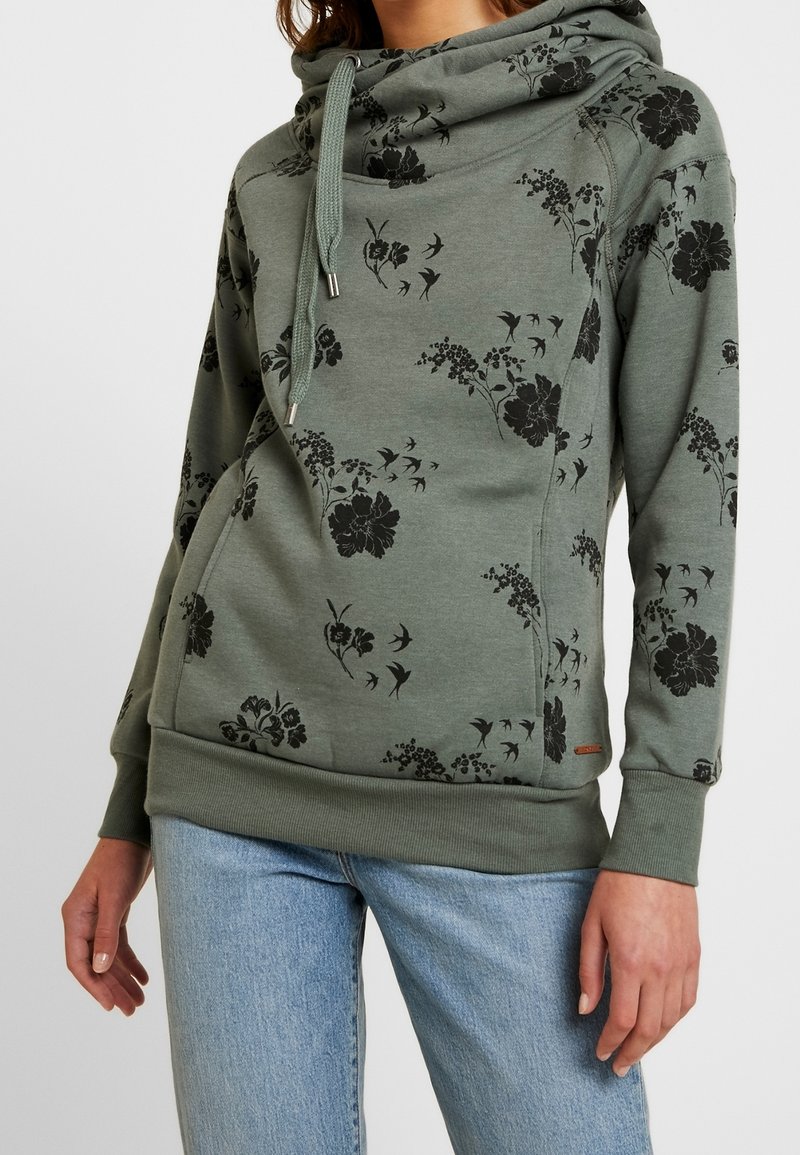 Femme portant un sweat à capuche vert avec un imprimé floral et oiseaux noir, associé à un jean bleu clair, debout devant un fond uni.