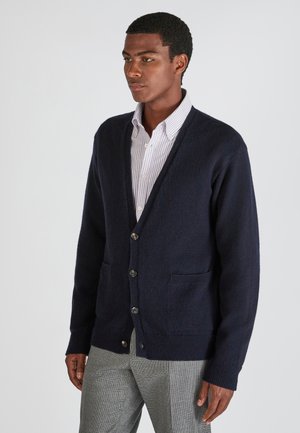 Marineblauer Strickcardigan mit V-Ausschnitt, Knopfverschluss und zwei Fronttaschen; strukturierter Stoff mit gerippten Bündchen und Saum. Über einem gestreiften Hemd getragen.