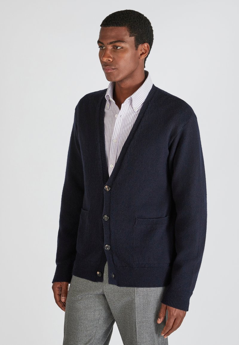 Cardigan en tricot marine avec décolleté en V, fermeture à boutons et deux poches avant ; tissu texturé avec poignets et ourlet côtelés. Porté par-dessus une chemise rayée.