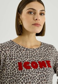 Camiseta con estampado de leopardo, cuello redondo y la palabra "ICONE" en letras rojas y llamativas. Tejido suave con un corte holgado y textura lisa.