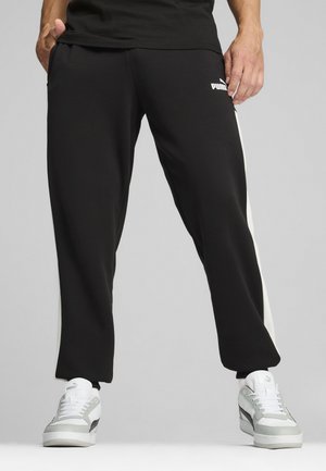Czarne spodnie sportowe z białymi paskami po bokach, o luźnym kroju i elastycznym pasie. Logo PUMA na lewej udzie. Noszone z białymi i szarymi trampkami.