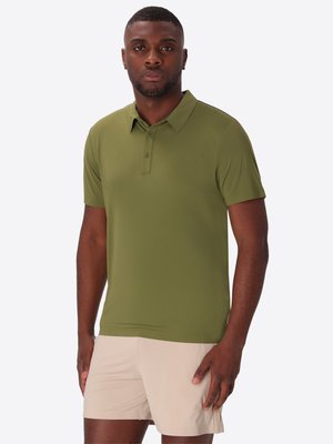 Uomo che indossa una polo verde oliva a maniche corte e pantaloncini beige chiaro, in piedi con un'espressione neutra su uno sfondo semplice.