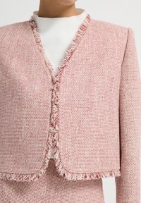 Chaqueta de tweed texturizada en rosa y blanco con bordes deshilachados, abertura frontal y un ajuste estructurado. Superpuesta sobre una blusa blanca con cuello alto.