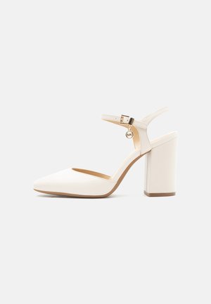 Schuh mit cremefarbenem Blockabsatz, spitzer Zehenpartie und Knöchelriemen, der einen kleinen goldenen Anhänger und eine Schnallenverschlus enthält.