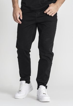 Homme portant un jean noir et des baskets blanches, debout les mains près de la taille devant un fond uni.