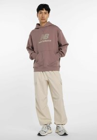 Sudadera de color marrón claro con un bolsillo frontal y logo, combinada con pantalones cargo crema que tienen bolsillos laterales, y zapatillas multicolores.