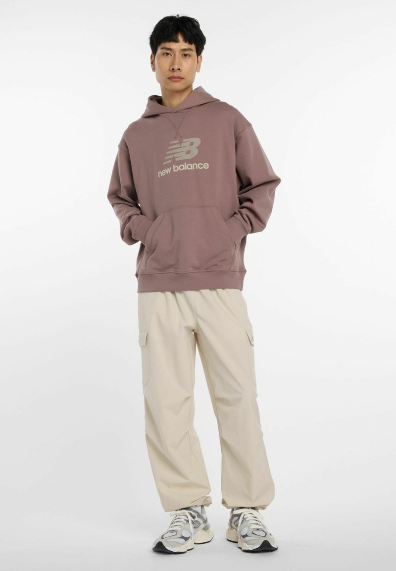 Sudadera de color marrón claro con un bolsillo frontal y logo, combinada con pantalones cargo crema que tienen bolsillos laterales, y zapatillas multicolores.