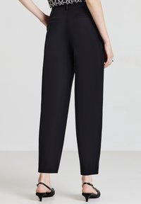 Pantalons noirs ajustés à coupe droite, avec poches arrière et texture lisse. Portés avec des talons noirs à brides.