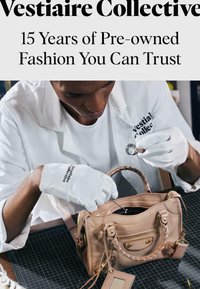 Personne portant des gants blancs inspectant un sac à main en cuir beige à l'aide d'une loupe sur un tapis à grille noire.