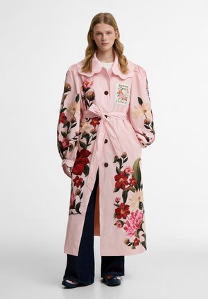 Naine kannab pikka roosat lillemustriga trentškotiga, millel on padrunvarrukad, krae, ees nupud, vöö vöökohal ja tumedad laiade säärtega püksid.