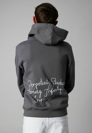 Asmuo dėvi tamsiai pilką džemperį su gaubtu, kuris uždėtas ant galvos, o ant nugaros baltomis ranka rašytomis raidėmis parašyta „Perpetuity Studio, Eternity Infinity Forever.“