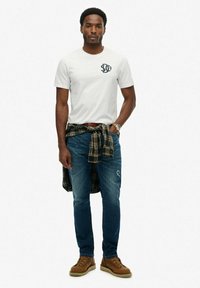 Hvid kortærmet t-shirt med et logo, blå jeans og brune snørebodstøvler; ternet skjorte bundet om livet. Afslappet look med minimale detaljer.