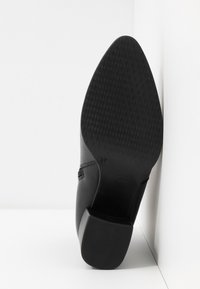 Bottine en cuir noir posée sur son talon, montrant la semelle avec un motif de crampons en forme de losange et la taille 37 marquée.