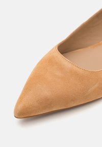 Sapato de camurça de bico fino em cor bege claro com textura suave e abertura em forma de amêndoa, apresentando um design minimalista e sem hardware visível.