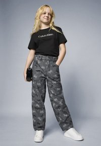 Must musta t-särk "Calvin Klein" logo ja hallid mustrilised ülepüksid, millel on valged tennisetossud. Mudel hoiab musta käekotti.
