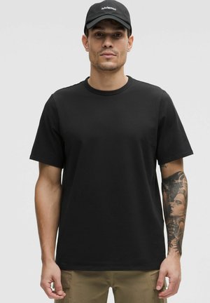 T-shirt nera a maniche corte con colletto rotondo, realizzata in cotone, con texture liscia. Indossata con pantaloni cargo verde oliva, che mostrano tatuaggi sulle braccia.