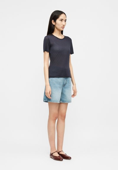 Jonge vrouw in zijaanzicht, gekleed in een donkergrijze nauwsluitende T-shirt, lichtblauwe denim shorts en donkerrode Mary Jane ballerina's.