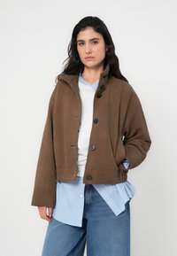 Καφέ κοντό μπουφάν με λεία υφή, που διαθέτει ψηλό γιακά, κουμπιά μπροστά και ραμμένους ώμους, φορεμένο πάνω από πουκάμισο με μπλε ρίγες.