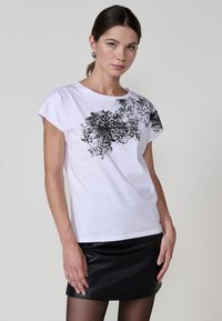 Camiseta blanca de algodón con un estampado abstracto negro en un lado. Presenta mangas cortas y un corte relajado, combinada con una falda negra texturizada.