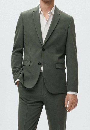 Uomo che indossa un abito verde oliva con due bottoni neri e una camicia bianca, con una mano in tasca, in piedi davanti a uno sfondo semplice.