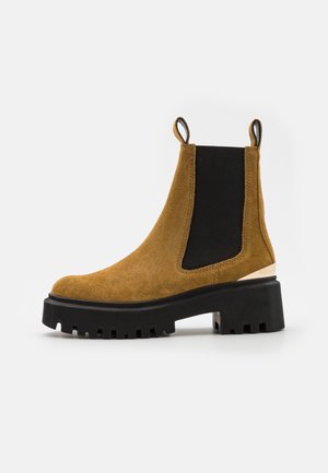 Brązowy zamszowy botek za kostkę z czarnymi elastycznymi panelami bocznymi, grubą czarną gumową podeszwą i pętlami do łatwego zakładania. Charakteryzuje się nowoczesnym designem.