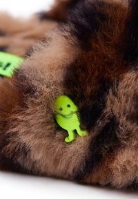 Material esponjoso con un patrón marrón y negro, con una pequeña figura de plástico verde incrustada, que presenta una sonrisa y detalles en las extremidades.