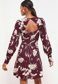 Vrouw met donker haar in een lage paardenstaart, draag een bordeauxrode bloemige jurk met lange mouwen en een driehoekige open rugdetail.