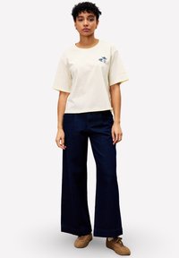 Vrouw met kort krullend haar, gekleed in een off-white T-shirt met een vlinderprint, donkere wijde broek en bruine casual schoenen, staat stil.