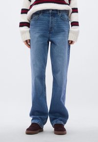 Φωτεινό γαλάζιο τζιν με άνετη εφαρμογή, σχεδιασμό με πέντε τσέπες, ορειχάλκινα εξαρτήματα και διακριτική ξεθώριαση. Φοριέται με σκουροκόκκινα αθλητικά παπούτσια.